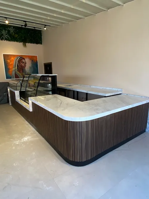 Koyu ahşap oluklu panelli, geniş beyaz mermer tablalı, L tipi modern cafe kasa bankosu ve pasta teşhir tezgahı