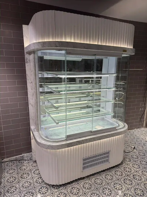 Oval hatlara sahip, beyaz oluklu panelli ve mermer tablalı, lüks dikey pastane teşhir reyonu ve soğutma vitrini